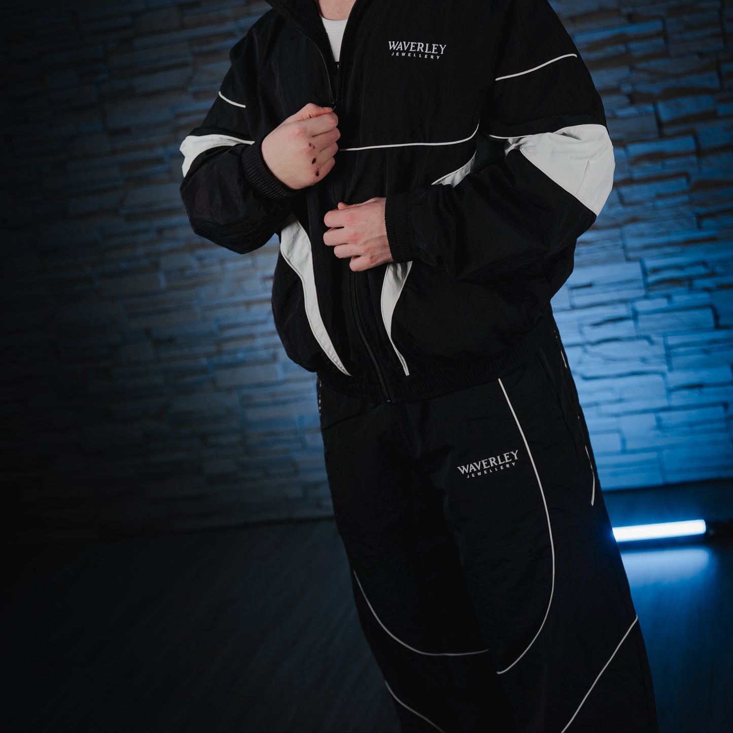Man wearing a black and white jacket with a logo against a dark brick wall.