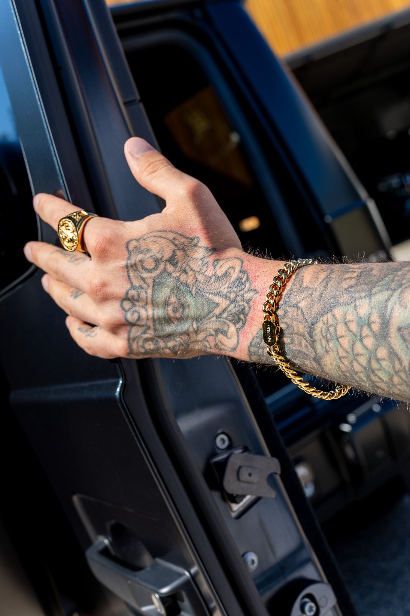 Hand with tattoos opening a car door, wearing gold rings and bracelets.