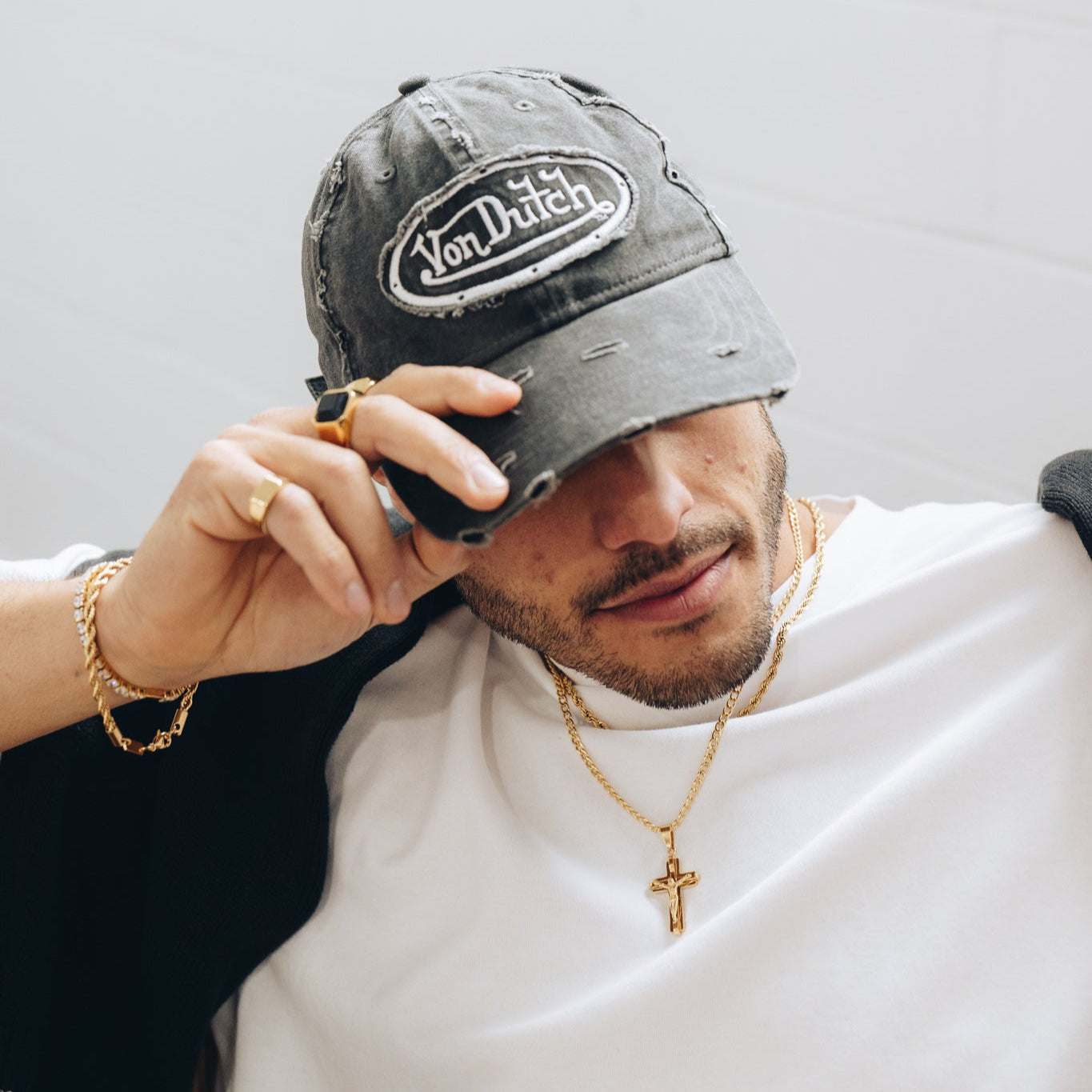 Man wearing a gray cap with a brand logo, holding a phone, against a plain background and gold chain