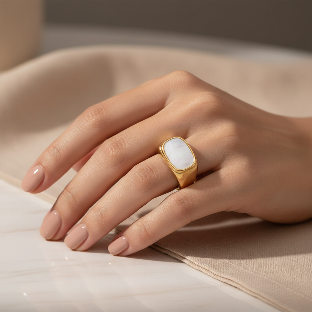 A close-up of a hand wearing a gold ring with a large, smooth white oval stone, resting on a beige fabric surface.